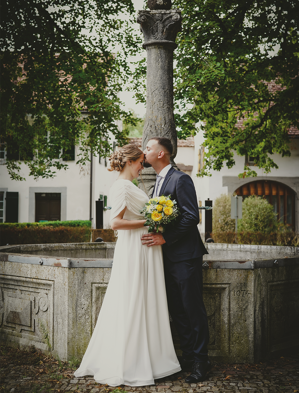 Brautpaar küsst sich liebevoll an historischem Brunnen.